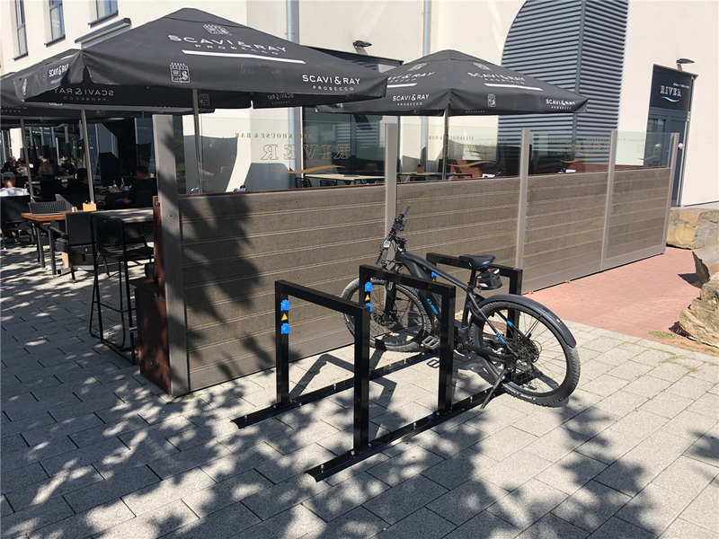 E-Bike Anlehnbügel ohne Ladeeinheit vor einem Restaurant mit Sonnenschirmen, Fahrrad sicher abgestellt auf gepflasterter Fläche