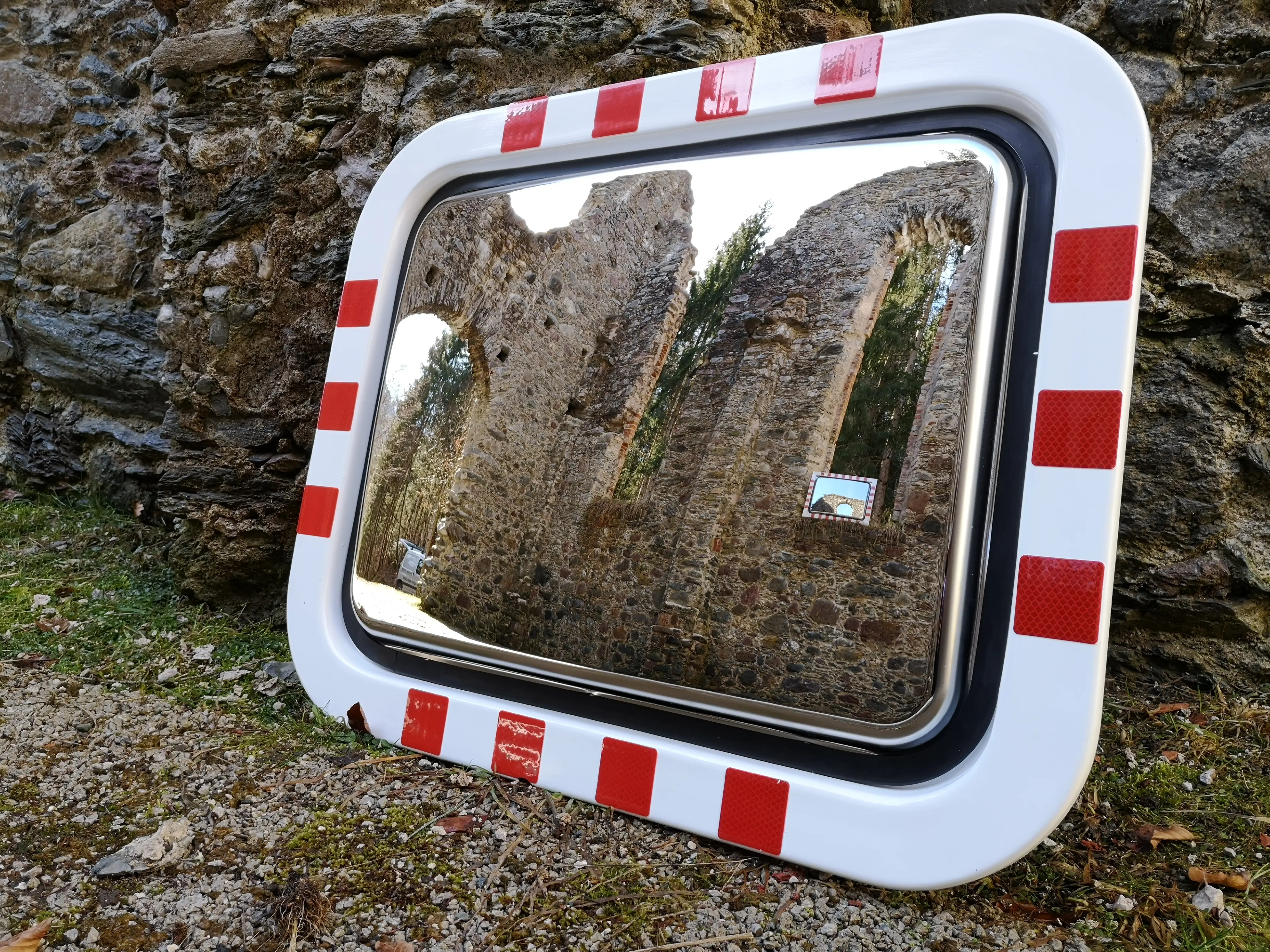 Verkehrsspiegel mit rot-weißem Rand lehnt an einer alten Steinmauer. In der Spiegelfläche spiegelt sich eine historische Ruine mit Bäumen im Hintergrund.