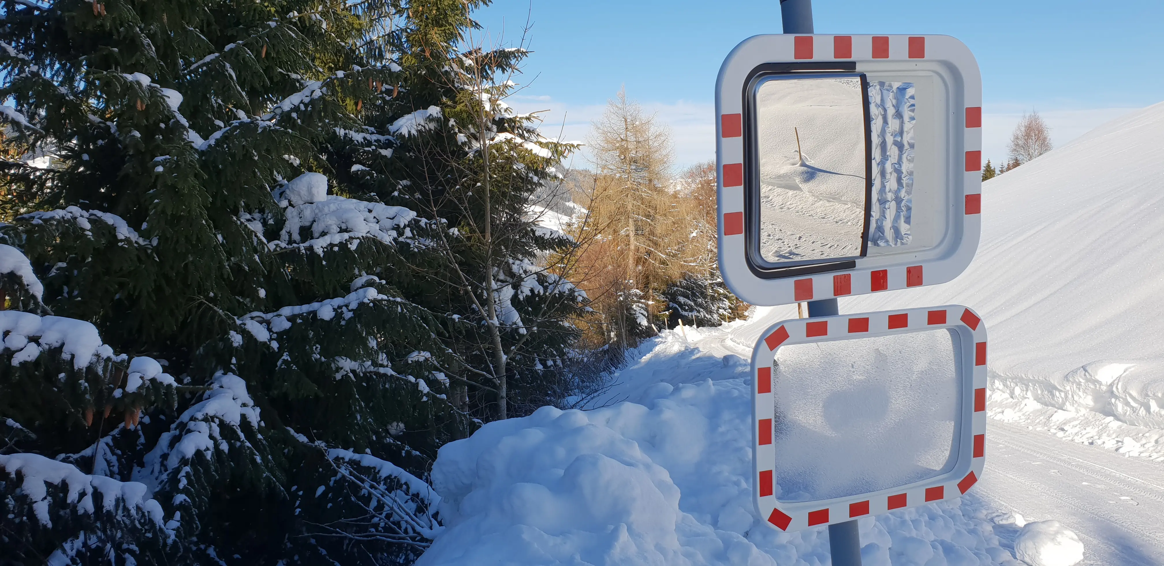 Zwei rechteckige Verkehrsspiegel mit rot-weißem Rahmen an einem Pfosten montiert, im winterlichen Gebirge. Der obere Spiegel zeigt eine klare Sicht auf einen Schneehang, während der untere Spiegel von Frost und Eis beschlagen ist.