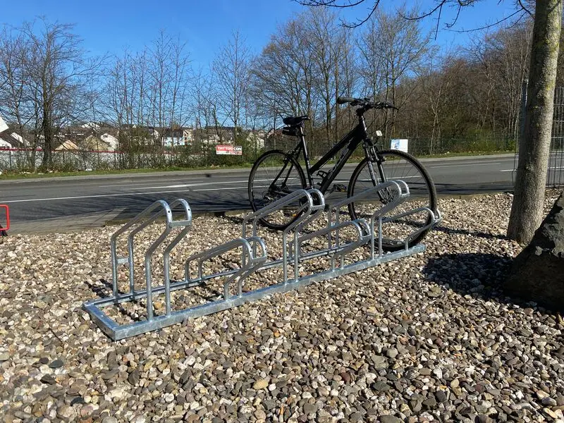  Fahrradständer mit sechs geraden Stellplätzen auf Schotterfläche, ein Fahrrad ist im Halter geparkt, Hintergrund mit Straße und Bäumen