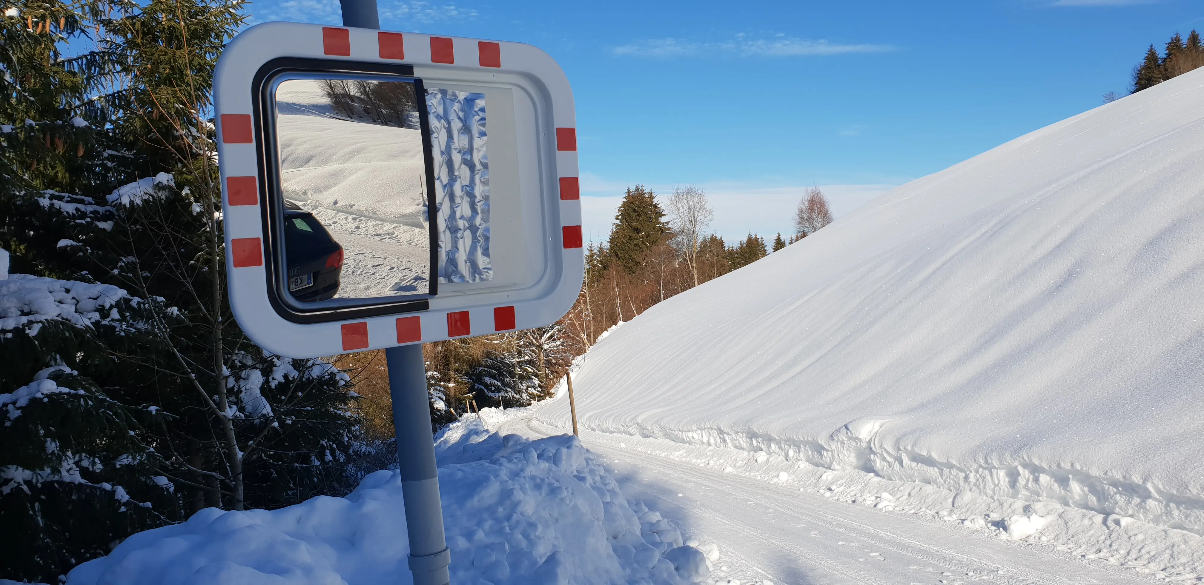 Verkehrsspiegel mit rot-weißer Umrandung an verschneiter Landstraße, spiegelt Fahrzeug und Winterlandschaft