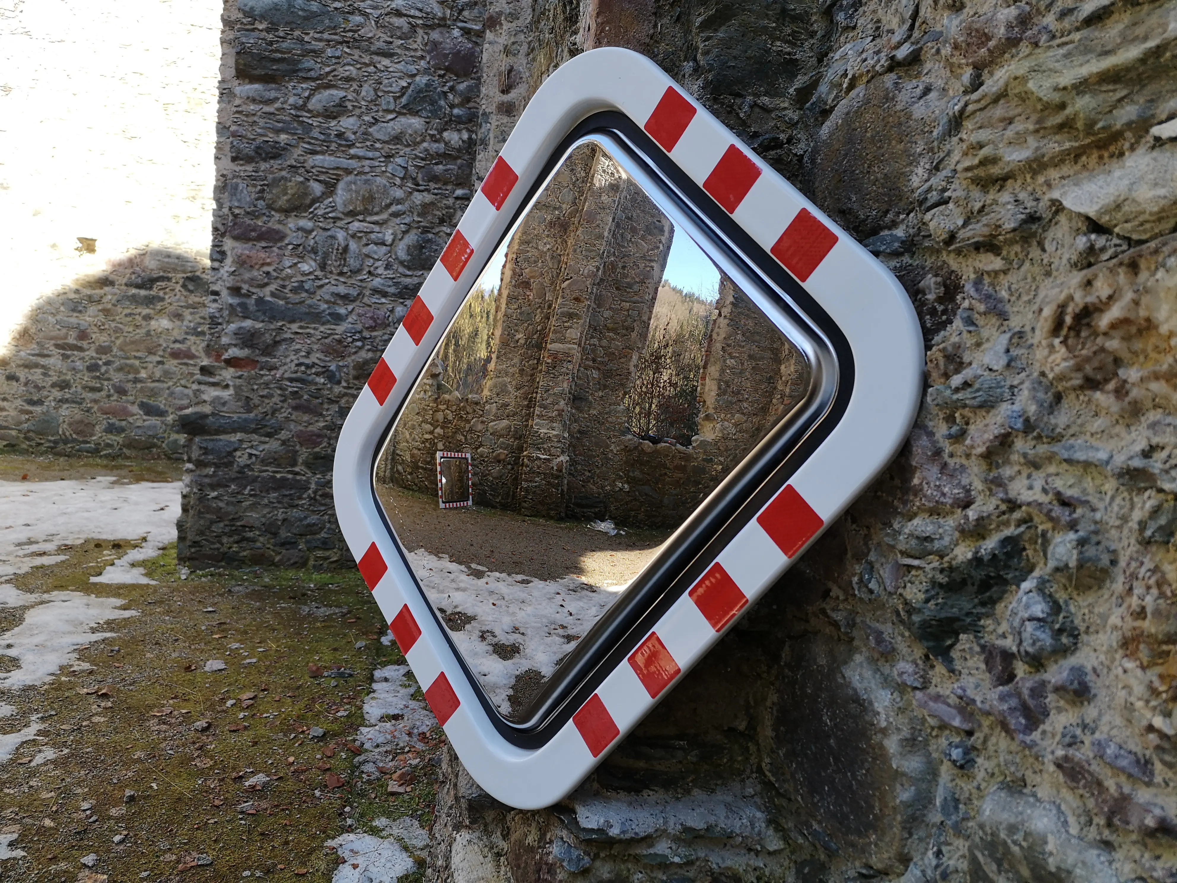 Verkehrsspiegel mit rot-weißer Markierung, diagonal montiert an einer Steinmauer; reflektiert eine historische Ruinenlandschaft mit einem weiteren Spiegel im Hintergrund.