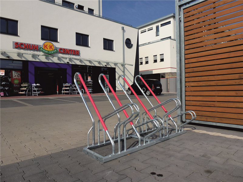 Fahrradständer mit roten Anlehnbügeln vor einem Geschäftszentrum auf gepflasterter Fläche montiert, bietet sichere Abstellplätze für mehrere Fahrräder